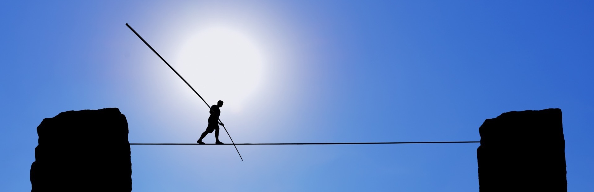 A man walking on a tightrope between two pillars of rock against a bright blue sky. He's using a long pole to help with his balance.